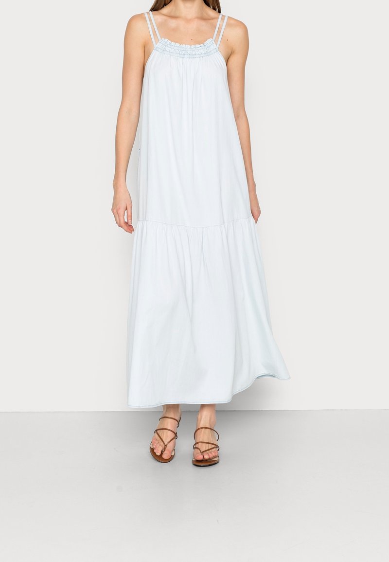 Femme portant une robe longue bleu clair sans manches avec des fines bretelles doubles et des sandales marron à lanières.