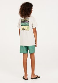 Lichtbeige T-shirt met een kleurrijk grafisch ontwerp op de achterkant, gecombineerd met groene shorts met een flespatroon en zwarte flip-flops.