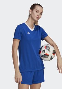 Blue athletic shirt with white V-neck and matching shorts. The player holds a soccer ball featuring black and red patterns, branded Adidas.