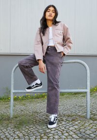 Kobieta opierająca się o metalową balustradę na zewnątrz, ubrana w fioletową kurtkę, biały top, szare spodnie, białe skarpetki i czarno-białe sneakersy.