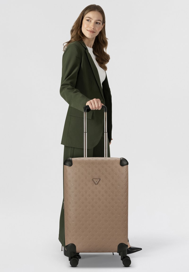 Mujer con traje verde oliva sosteniendo el mango de una maleta rodante beige con un patrón y cuatro ruedas, de pie contra un fondo blanco.