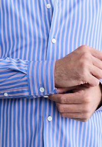 Plava košulja na kopčanje s uskim vertikalnim crvenim i bijelim prugama. Ima klasični ovratnik i manžete s bijelim gumbima. Glatka tekstura tkanine.