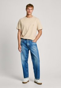 Noor mees kannab beeži t-särki ja siniseid teksaseid, millel on väike rebend. Ta seisab ühe käega taskus ja jalas on tal valged tossud, taust on ühtlane.