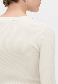 Beige ribbed knit top with a round neckline, featuring a subtle shimmer and fitted sleeves. Back view highlights texture and seam details.