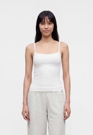 Jeune femme aux longs cheveux foncés, portant un débardeur blanc et un pantalon ample gris clair, debout devant un fond uni.