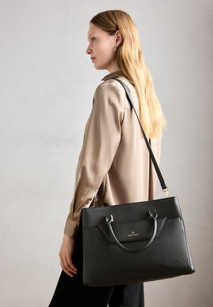 Woman with long blonde hair wears beige blouse and black pants, carrying a large black leather handbag with shoulder strap.