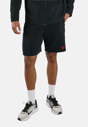 Man draagt zwarte Reebok shorts, witte kousen en zwart-witte Reebok sneakers en staat tegen een witte achtergrond.