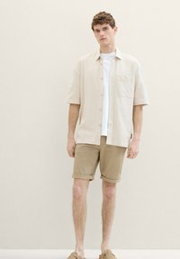 Cămașă cu mânecă scurtă, de culoare bej deschis, cu butoni, având un buzunar pe piept, purtată peste un tricou alb, asociată cu pantaloni scurți khaki răsuciți și pantofi de culoare brun deschis.