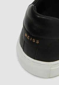 Fekete bőrsneaker struktúrált felső résszel és fehér gumitalppal. Az arany színű "REISS" márkázás a sarkon egy diszkrét részletet ad.