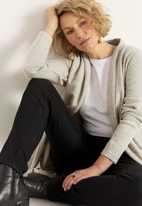 Beige ribbed cardigan ylle valkoisen paidan päällä, mustien housujen kanssa. Yhdistetty mustiin nilkkureihin ja suuriin näyttäviin korvakoruihin.