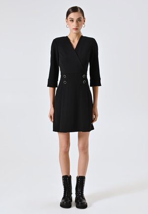 Schwarzes Wickelkleid mit Dreiviertelärmeln, das doppelte Kreisakzente an der Taille aufweist. Gestylt mit klobigen schwarzen Schnürstiefeln.