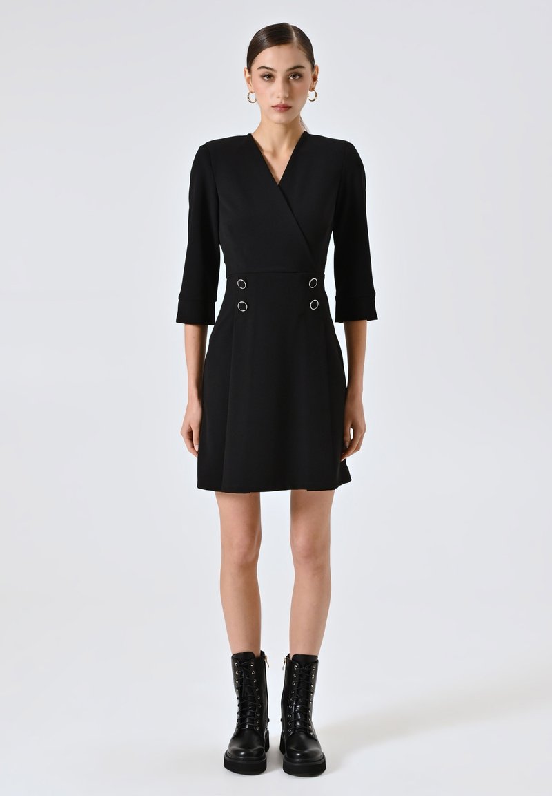 Schwarzes Wickelkleid mit Dreiviertelärmeln, das doppelte Kreisakzente an der Taille aufweist. Gestylt mit klobigen schwarzen Schnürstiefeln.