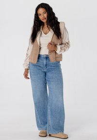 Beige vest met een donzige kraag en bloemenbroderie, gecombineerd met lichtblauwe wijde jeans en beige instappers op een witte achtergrond.