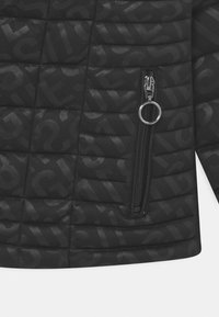 Section de veste matelassée noire avec un motif subtil de texte en all-over et une poche zippée verticale dotée d'un anneau de tirage en métal rond.