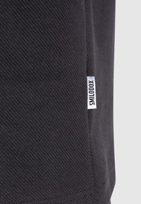 Gros plan d’un tissu noir texturé avec une petite étiquette rectangulaire blanche portant la mention "SMILODOX" cousue dans la couture latérale.