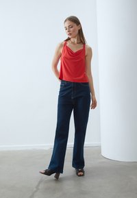 Top senza maniche rosso con scollo a cowl, abbinato a jeans a gamba larga blu scuro. Le scarpe sono nere con tacco a blocco e presentano un design con punta aperta.