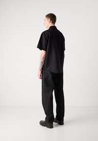 Camisa negra de manga corta con un ajuste relajado, combinada con pantalones de mezclilla oscuros que presentan un detalle de costura lateral y zapatos de cuero negros.