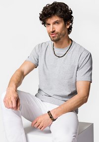 Szary bawełniany t-shirt z krótkimi rękawami i kieszonką na piersi. W połączeniu z białymi spodniami, ozdobiony biżuterią z koralików na szyi i nadgarstku.