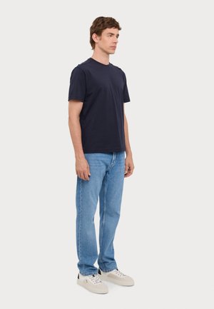 Camiseta de algodón azul marino de manga corta, combinada con jeans rectos de color azul claro y zapatillas blancas suaves, de pie contra un fondo blanco.