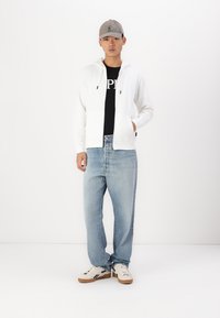 Witte zip-up hoodie over een zwart T-shirt, lichtblauwe jeans en beige sneakers met donkere accenten. Model draagt een grijze pet.