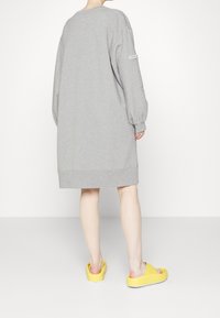 Grå oversized sweatshirtklänning med långa ärmar, ribbat nederkant och sidokutningar, i kombination med gula slip-on skor. Enfärgad bakgrund.