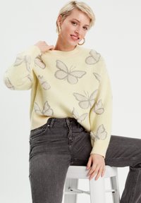 Gul strikket sweater med grå sommerfuglmønstre, rund halsudskæring og lange ærmer, parret med mørkegrå højtaljede jeans.