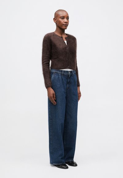Cárdigan marrón esponjoso con un escote redondeado y botones frontales, combinado con jeans oscuros de corte suelto y zapatos negros de punta.