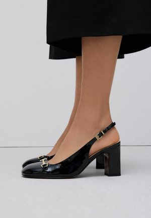 Jambes portant des escarpins slingback à talons blocs en cuir verni noir avec des détails de boucle dorée et un ourlet de jupe noir visible au-dessus des chevilles.