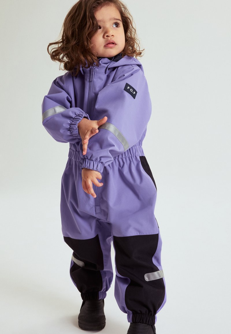 Peuter met krullend haar, gekleed in een paarse en zwarte waterdichte overall met reflecterende strips, kijkt omhoog en past een mouw aan.