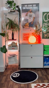 Rafturi din metal cu plante în ghiveci, un covor rotund negru cu un design de minge de biliard albă, un dulap gri și artă murală colorată cu o lampă orbitoare.