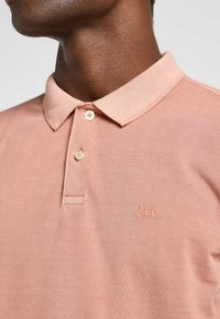 Ljusrosa polo skjorta gjord av texturerat tyg, med klassisk krage, tre knappar och en broderad 'Lee' logotyp på bröstet.
