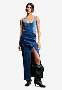 Rochie maxi din denim cu bust fix și despicătură laterală. Are un amestec de nuanțe de albastru închis și deschis, cu detalii strânse și o textură netedă.