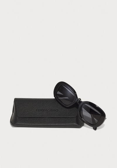 Černé oversized sluneční brýle s tmavými skly, ležící na černém texturovaném pouzdře s nápisem "Ferragamo". Pouzdro má elegantní, minimalistický design.