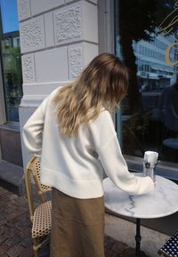 Krēmsvārti džemperis ar ribotu apakšmalu, saskaņots ar brūnu zamšādas svārku. Apaļs marmora galds ar traukiem turētājā blakus bistro krēslam.