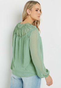 Blusa leggera di colore verde menta con pois, scollo smock e maniche arricciate. Presenta una vestibilità comoda e una texture trasparente.