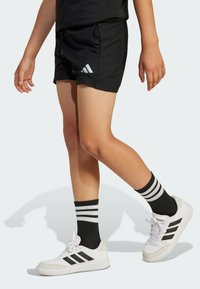 Čierne športové šortky so bočnými vreckami a elastickým pásom. V kombinácii s bielymi teniskami, čiernymi pruhovanými ponožkami, ktorých nositeľ zobrazuje dynamickú chôdzu.