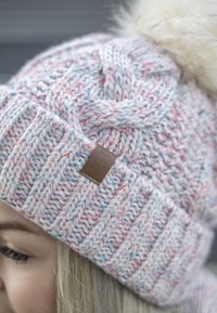 Strikket hue i pastelfarver med et fletmønster, prydet med en falsk pels pom-pom på toppen og et lille brunt lædermærke på siden.
