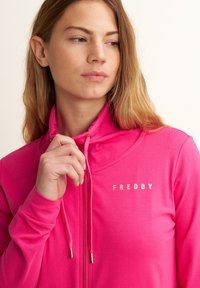 Veste zippée fuchsia en tissu doux, avec un col montant, des cordons de serrage et un logo doré "FREDDY" sur la poitrine.
