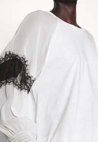 Top blanco con mangas abullonadas, con detalles de encaje negro en la manga izquierda y una tela texturizada y fruncida en el puño.