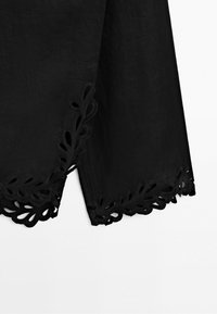 Tissu noir avec ourlet festonné et détail de broderie ajourée florale le long du bord, présenté sur un fond blanc uni.