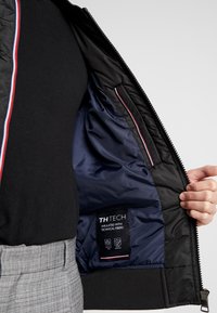 Sort isoleret jakke med quiltet ydre, marineblå foret og flerfarvet indvendig trim. Taggen læser "TH TECH, isoleret med tekniske fibre."