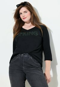 Schwarzes langärmliges T-Shirt mit V-Ausschnitt, mit grünen Strassbuchstaben, die "SPRING" sagen. Kombiniert mit dunkelgrauen Jeans.