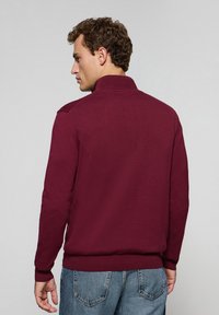 Suéter de punto burdeos con cuello alto, puños y dobladillo acanalados, textura suave y sutil detalle en los hombros.
