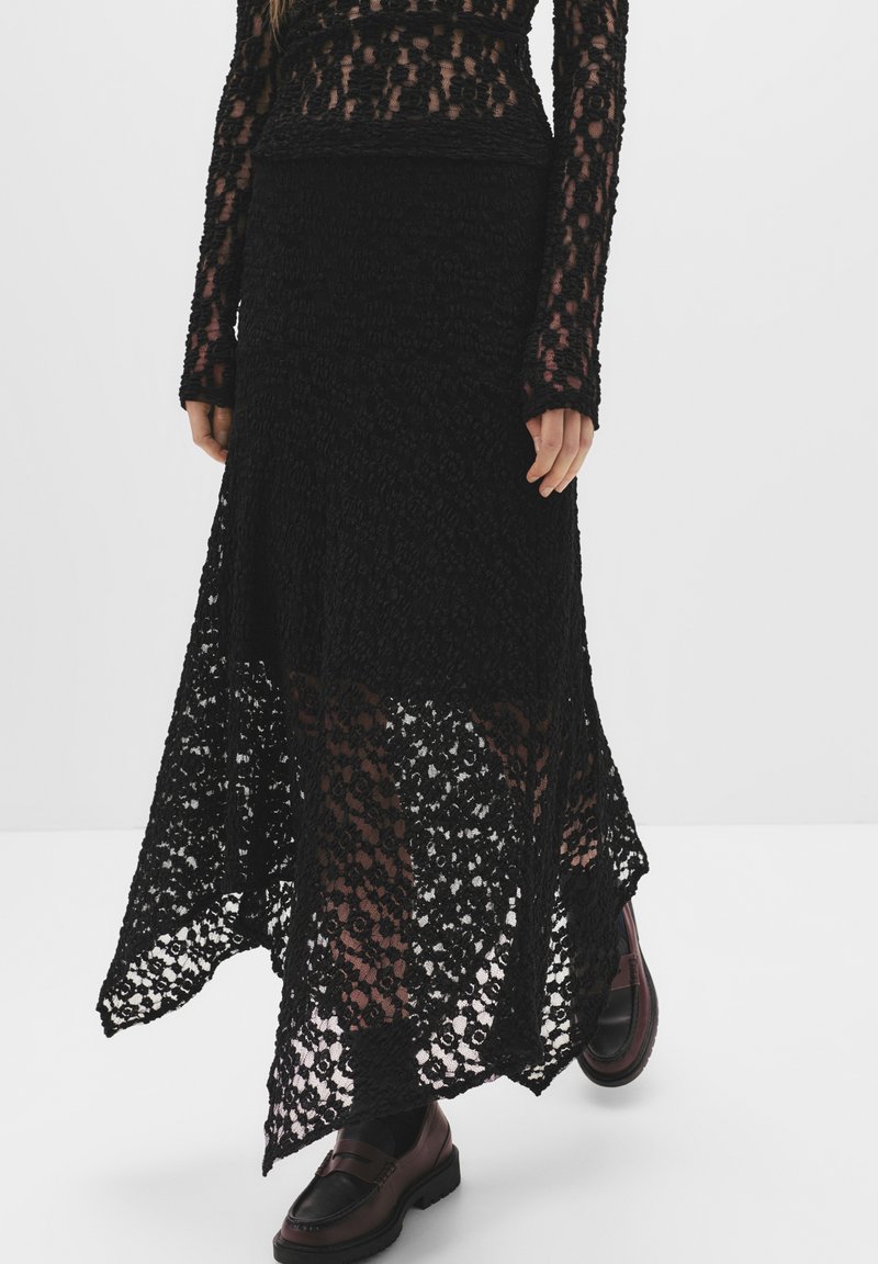 Gonna maxi in pizzo nero con un design fluido. Presenta sezioni trasparenti e un motivo testurizzato. Indossata con mocassini marroni scuri.