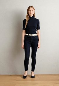 Haut à col roulé marine, jeans noirs slim, ceinture blanche et ballerines noires. Texture lisse, design ajusté, debout contre un mur clair.