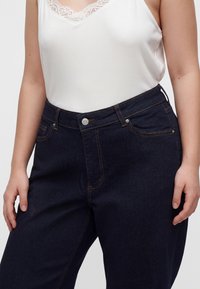Femme portant un jean taille haute bleu foncé et un top blanc sans manches avec bordure en dentelle, mains détendues le long du corps.