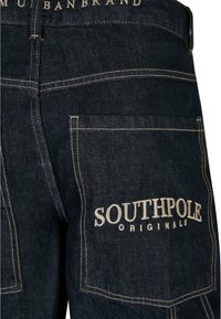 Tummansiniset farkut, joissa on näkyvät tikkaus yksityiskohdat, suurella takataskulla, jossa on valkoisella brodeerattu teksti "SOUTHPOLE ORIGINALS".
