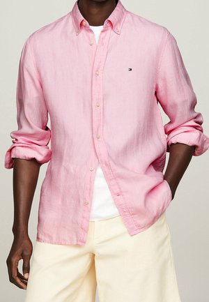 Osoba o ciemnej karnacji, ubrana w jasno-różową koszulę na guziki nałożoną na białą podkoszulkę oraz kremowe spodnie, z nonszalancko podwiniętymi rękawami.