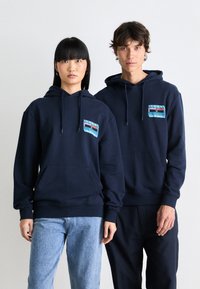 Hoodies bleu marine avec poches avant, capuches à cordon, et un patch graphique contrastant sur la poitrine. Les modèles portent des jeans décontractés et des joggings.