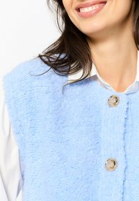 Gaiši zils teksturēts veste ar pūkainu audumu, ar zelta pogu akcentiem un baltu krekliņu ar apkaklīti zemāk.